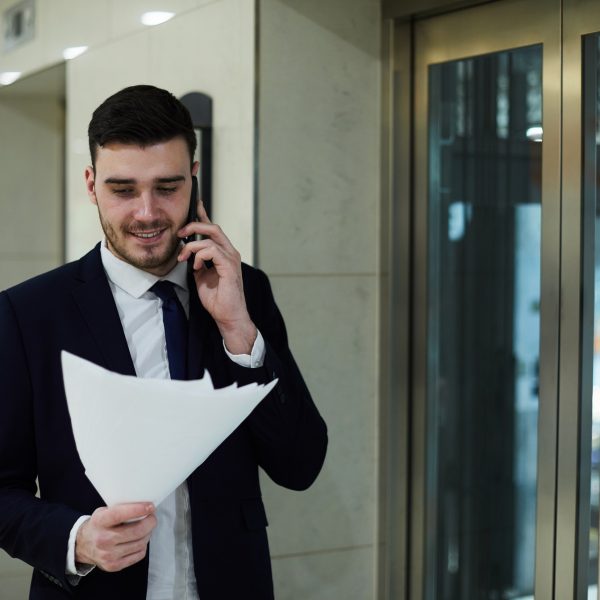 Cheerful confident handsome male manager in black suit walking by elevators and talking to business partner on phone while reading papers