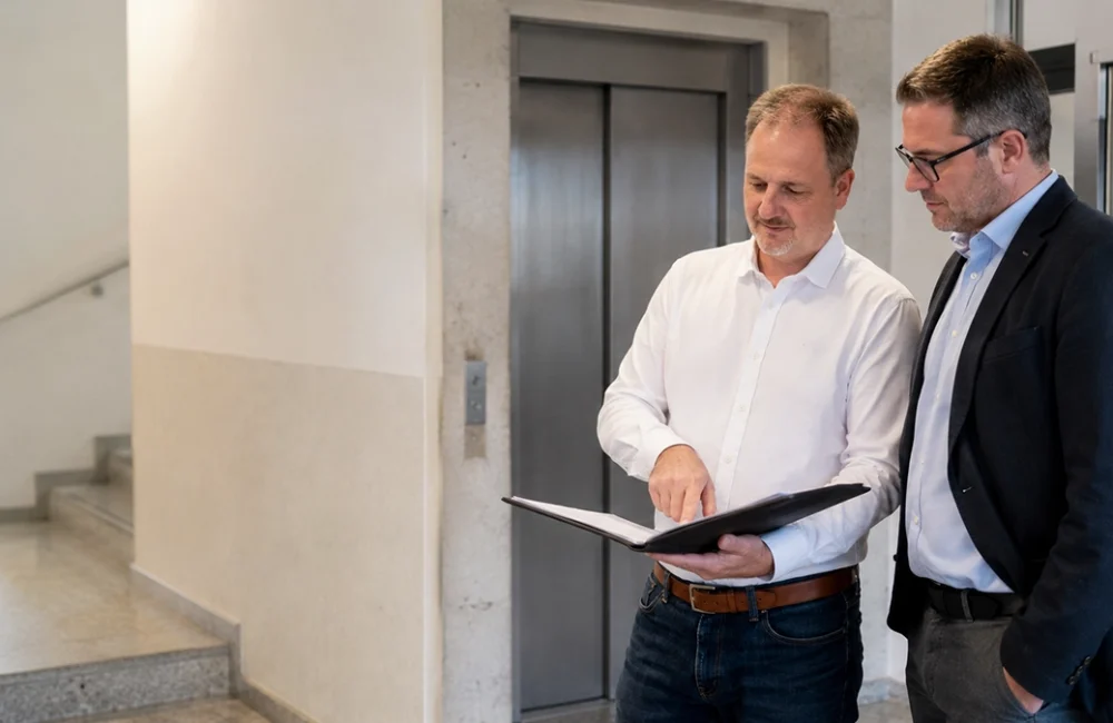 Zwei Männer besprechen Unterlagen vor einer Aufzugstür in einem Gebäudeflur
